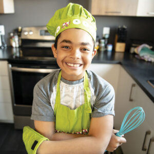 smiling boy wearing green bakers hat, apron and oven mitt, holding whisk