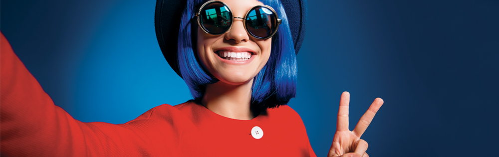 teen girl with blue hair and round sunglasses wears hat and red jacket. She smiles and does the peace sign.